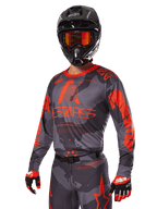 Zawodnik motocrossowy w zestawie Alpinestars 2025 Racer Hollow Red, szaro-czarny wzór kamuflażu z wyrazistym czerwonym brandingiem, komplet z koszulką, spodniami, rękawicami oraz czarnym kaskiem z czerwonymi goglami.