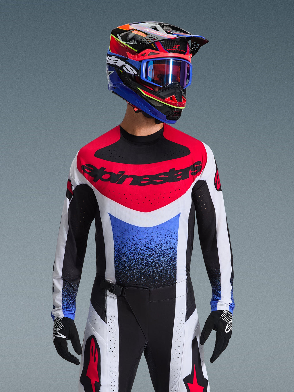 Koszulka Alpinestars 2026 Techstar Knif, koszulka motocrossowa, czerwono-biało-niebieska, z wyraźnym logo na klatce piersiowej i gradientem na tułowiu, noszona przez zawodnika w pasującym wielokolorowym kasku, niebieskich goglach i czarnych rękawicach, profesjonalna odzież wyścigowa off-road.