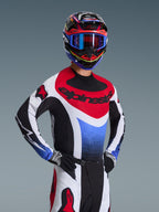 Motocyklista motocrossowy w koszulce Alpinestars 2026 Techstar Knif w barwach czerwono-biało-niebieskich, z czarnym brandingiem na klatce piersiowej, w zestawie z dopasowanymi spodniami motocrossowymi, pasującym kaskiem wyścigowym z niebieskimi goglami i czarno-białymi rękawicami, profesjonalny ubiór off-road do rywalizacji