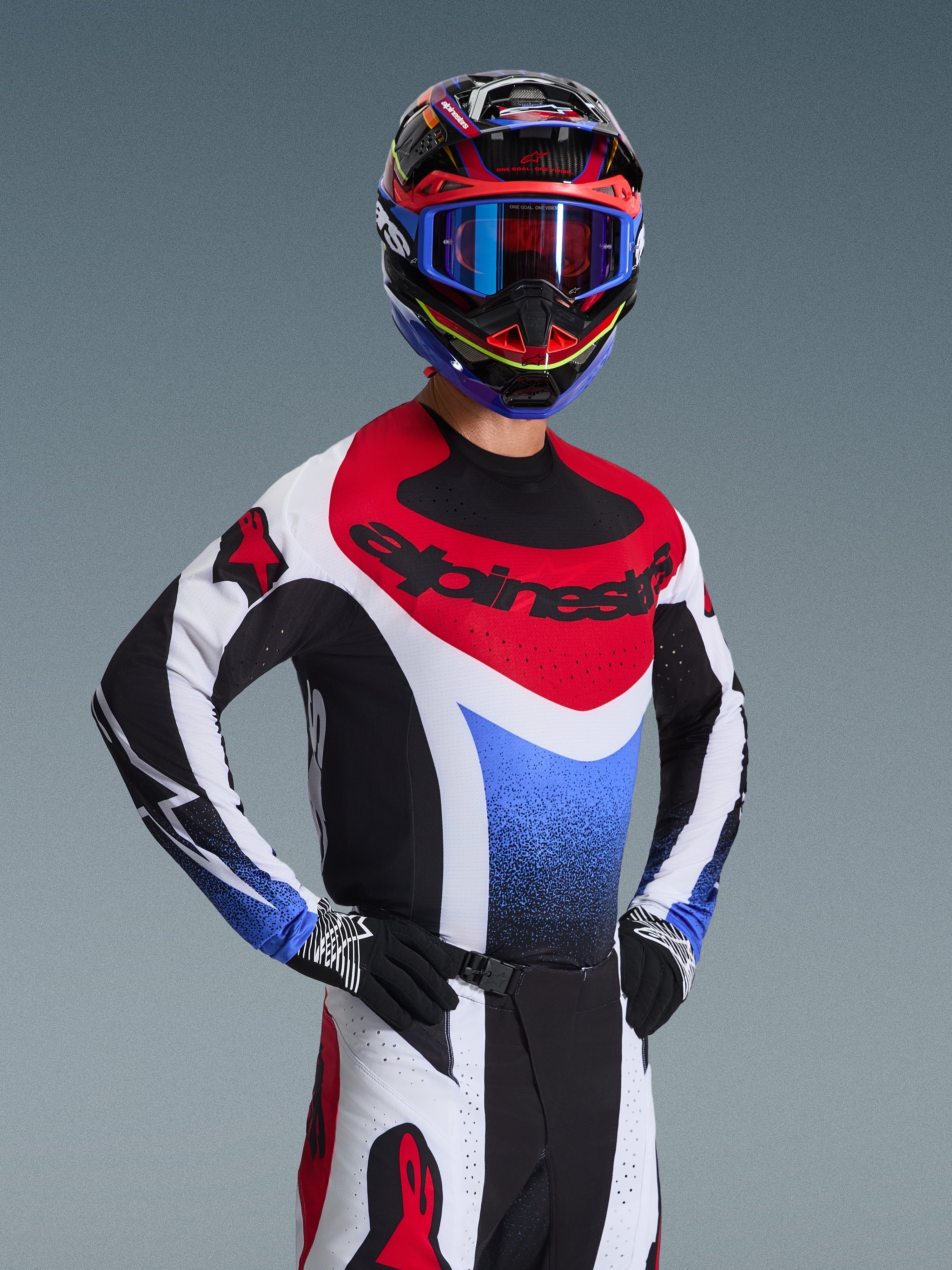 Motocyklista motocrossowy w koszulce Alpinestars 2026 Techstar Knif w barwach czerwono-biało-niebieskich, z czarnym brandingiem na klatce piersiowej, w zestawie z dopasowanymi spodniami motocrossowymi, pasującym kaskiem wyścigowym z niebieskimi goglami i czarno-białymi rękawicami, profesjonalny ubiór off-road do rywalizacji