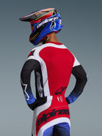 Zawodnik motocrossowy Alpinestars widziany od tyłu, koszulka 2026 Techstar Knif w kolorze czerwono-biało-czarnym, laserowo wycinane perforacje na plecach, wyrazisty branding, dopasowane spodnie z niebieskimi akcentami, niebiesko-czarny kask, profesjonalna estetyka off-road