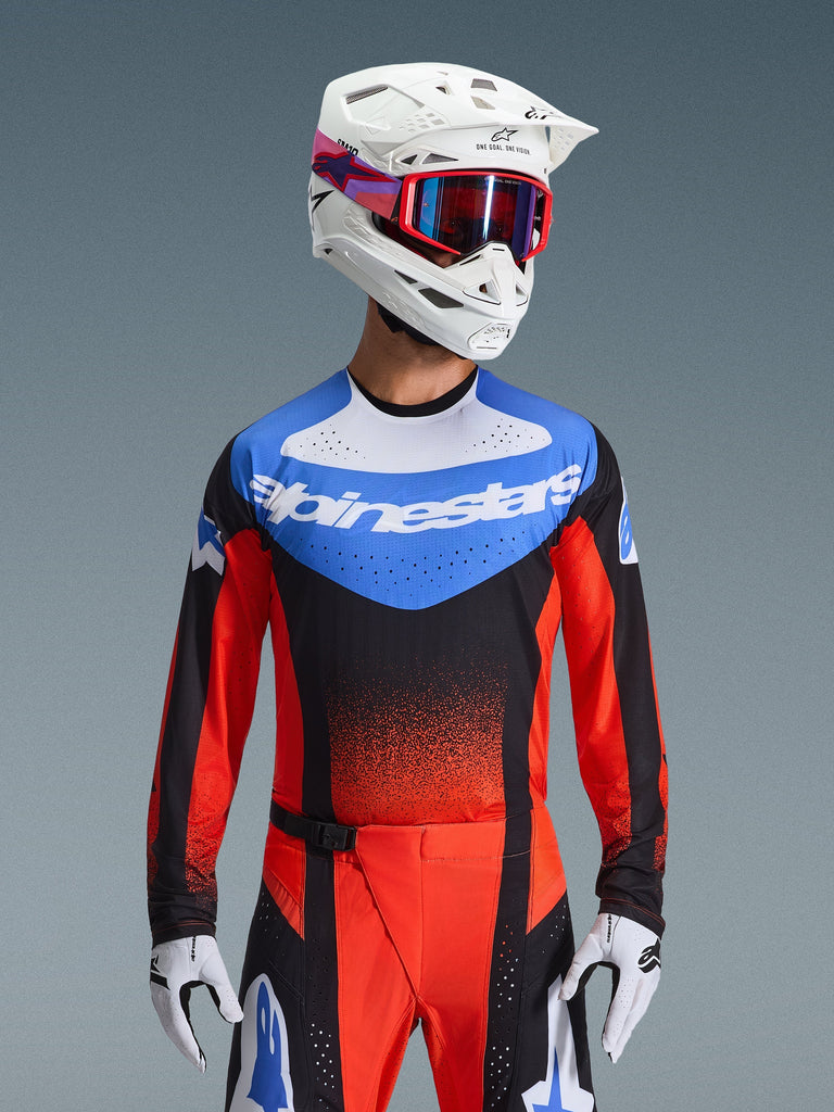 Alpinestars 2026 Techstar Knif Jersey, koszulka motocrossowa, kolor pomarańczowo-czarny i błękit UCLA Blue, posiada niebieski panel na klatce piersiowej z białym logo Alpinestars i pomarańczowo-czerwone rękawy, zestawiona z białym kaskiem, czerwonymi goglami i białymi rękawicami, profesjonalna odzież do wyścigów off-road