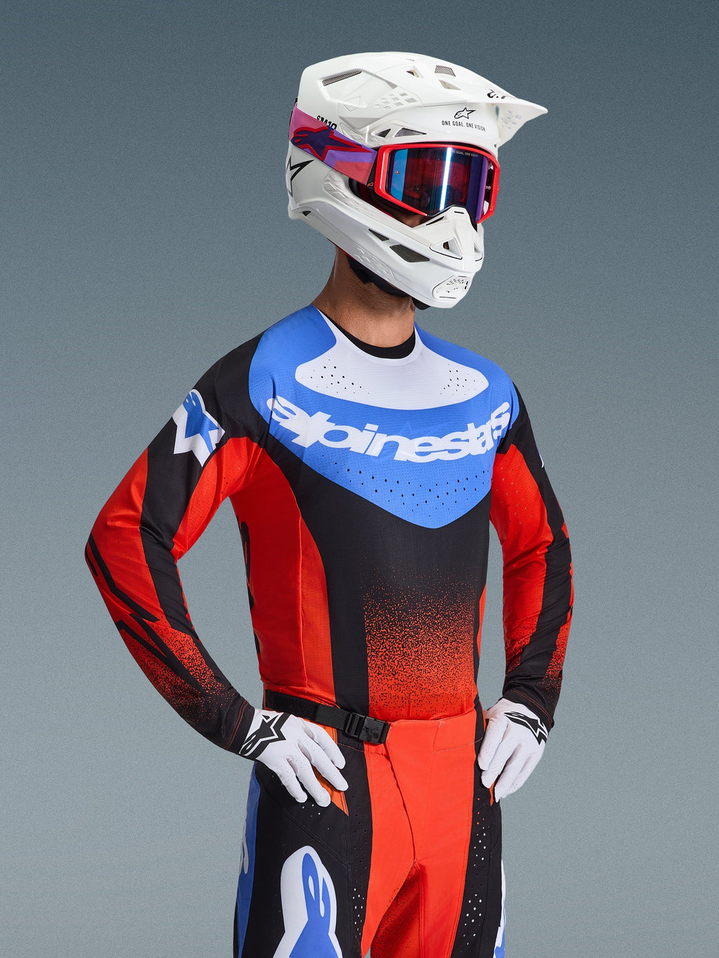Zawodnik motocrossowy ubrany w koszulkę Techstar Knif 2026 w kolorach pomarańczowym, czarnym i UCLA Blue, z niebieskim panelem na klatce piersiowej i białym logo Alpinestars, czarno-pomarańczowymi rękawami z laserowymi perforacjami, w zestawieniu z białym kaskiem, czerwonymi goglami i dopasowanymi spodniami wyścigowymi.