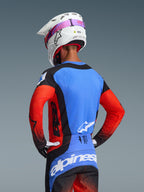 Motocyklista motocrossowy widziany od tyłu w zestawie Alpinestars Techstar Knif 2026, niebieska koszulka z czerwonymi rękawami i czarnymi akcentami, czarno-niebieskie spodnie z białym logo Alpinestars, biały kask z różowym paskiem gogli, z laserowo wyciętymi otworami wentylacyjnymi