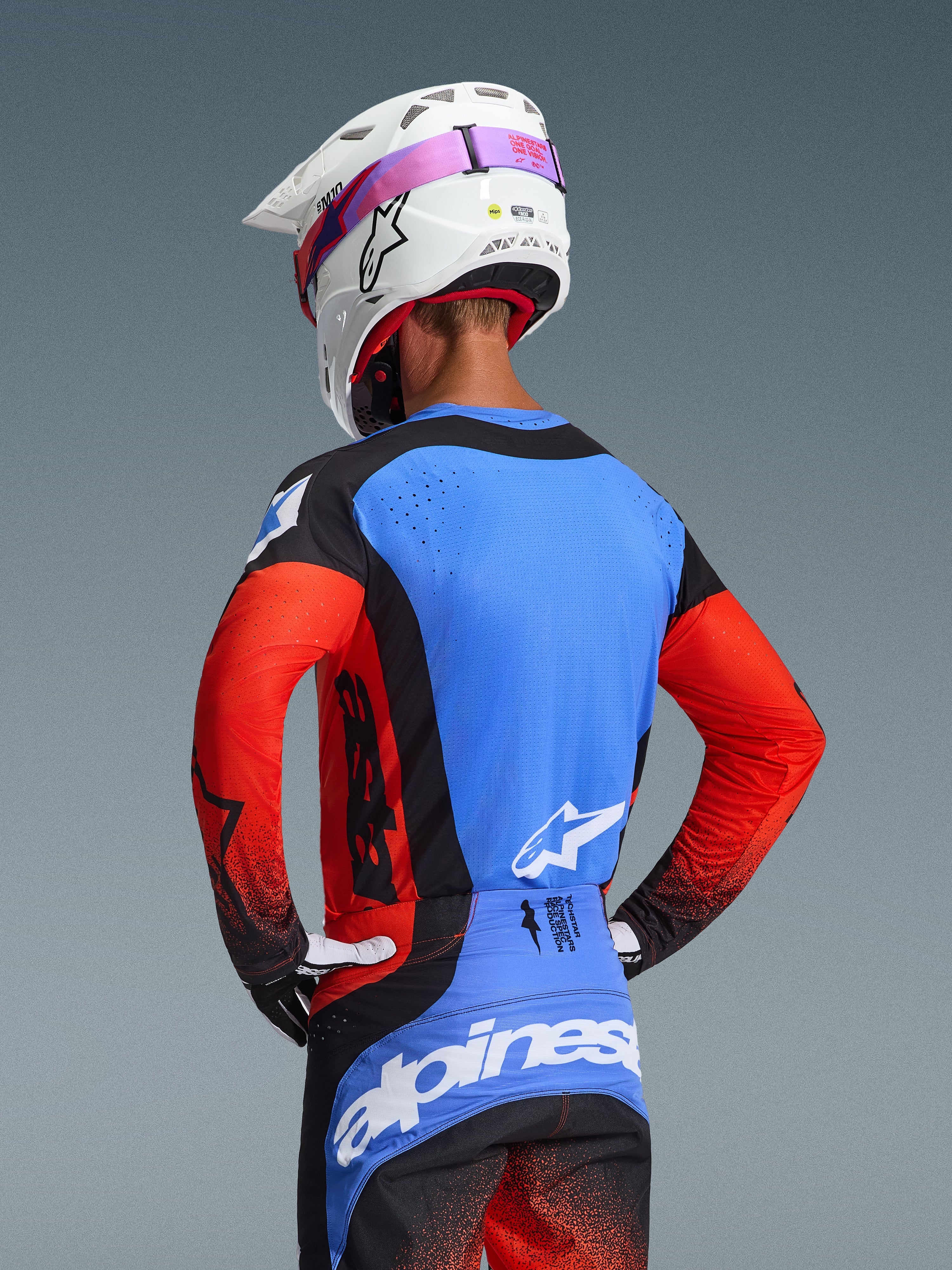 Motocyklista motocrossowy widziany od tyłu w zestawie Alpinestars Techstar Knif 2026, niebieska koszulka z czerwonymi rękawami i czarnymi akcentami, czarno-niebieskie spodnie z białym logo Alpinestars, biały kask z różowym paskiem gogli, z laserowo wyciętymi otworami wentylacyjnymi
