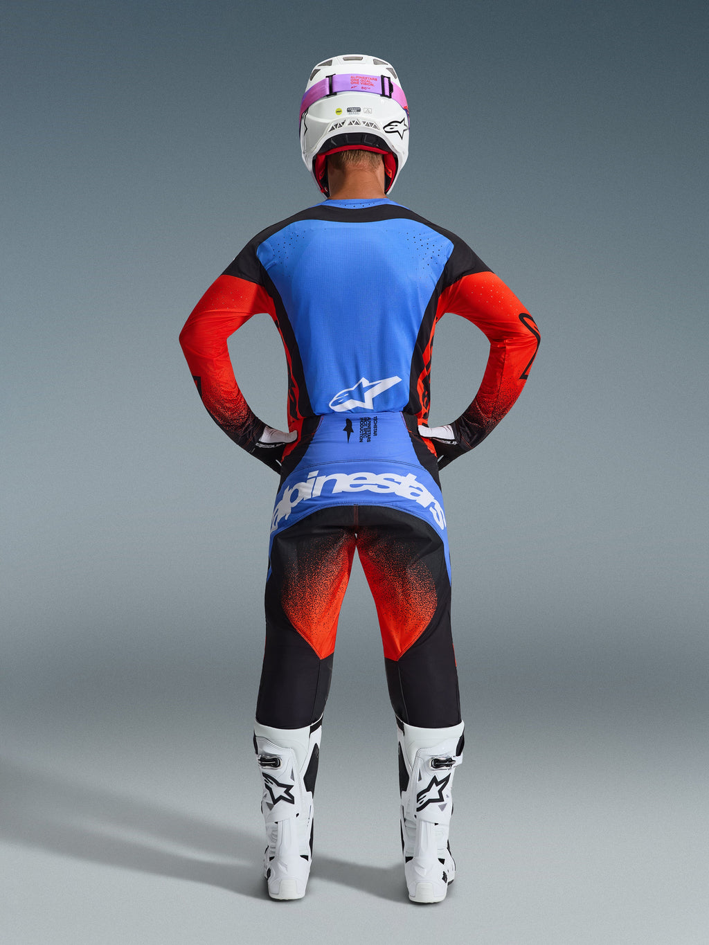 Widok od tyłu motocyklisty motocrossowego w zestawie Alpinestars 2026 Techstar Knif, niebiesko-czerwona koszulka z czarnymi akcentami, czarne spodnie z czerwonym gradientem na udach i niebieskim brandingiem, białe buty wyścigowe i biały kask z różowymi goglami.
