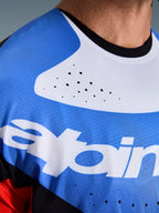 Alpinestars 2026 Techstar Knif Jersey, koszulka motocrossowa, zbliżenie na klatkę piersiową i szyję, niebiesko-biała z czarnym kołnierzykiem, laserowo wycinane otwory wentylacyjne, oddychająca siateczkowa tkanina z logo Alpinestars