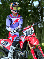 Zawodnik motocrossowy na czerwonym motocyklu Honda CRF, koszulka Alpinestars 2025 Fluid Wurx fioletowo-jaskrawoczerwona, pasujące fioletowo-czerwone spodnie, kask Red Bull z żółtymi goglami, białe buty i rękawice Alpinestars, profesjonalny sprzęt MX do jazdy terenowej