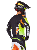 Motocyklista motocrossowy widziany od tyłu w zestawie Alpinestars 2025 Fluid Wurx, czarna koszulka z wyrazistymi neonowo-żółtymi i pomarańczowymi grafikami, dopasowane spodnie z białymi i pomarańczowymi panelami, czarny kask z neonowymi akcentami, wyraźny branding Alpinestars na rękawach i siedzeniu