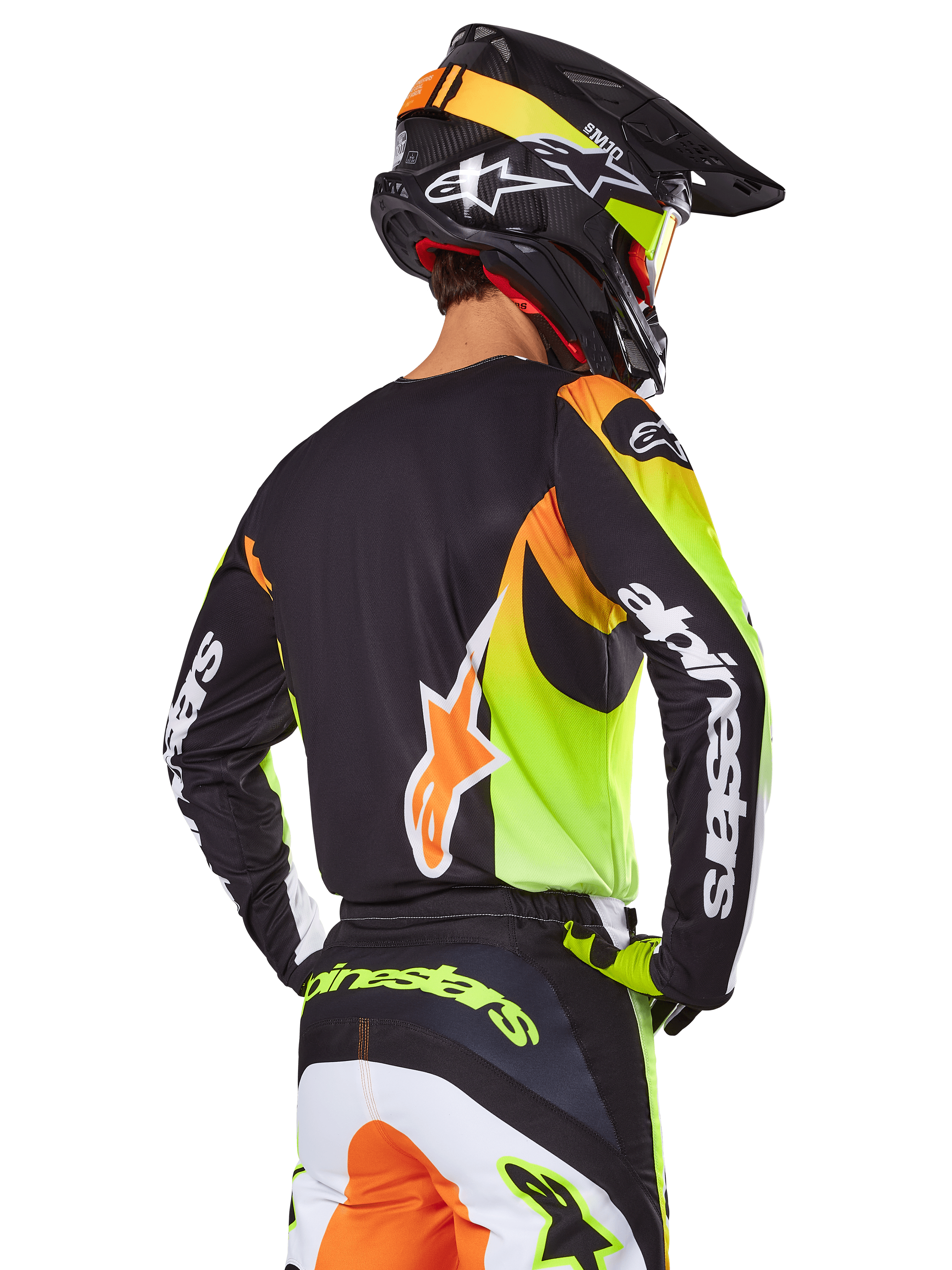 Motocyklista motocrossowy widziany od tyłu w zestawie Alpinestars 2025 Fluid Wurx, czarna koszulka z wyrazistymi neonowo-żółtymi i pomarańczowymi grafikami, dopasowane spodnie z białymi i pomarańczowymi panelami, czarny kask z neonowymi akcentami, wyraźny branding Alpinestars na rękawach i siedzeniu