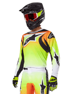 Motocyklista motocrossowy w zestawie Alpinestars 2025 Fluid Wurx, koszulka z żółto-pomarańczowym gradientem, czarnymi akcentami i dużym logo gwiazdy, pasujące spodnie motocrossowe, czarny kask z czerwonymi goglami i żółte rękawice, skoordynowany wyczynowy strój do wyścigów terenowych