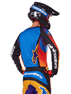 Motocrossowiec widziany od tyłu w zestawie Alpinestars 2025 Fluid Wurx, wielokolorowa koszulka z niebieskim tyłem i czarnymi rękawami z białym logo, pasujące pomarańczowo-niebieskie spodnie, czarno-niebieski kask z pomarańczowymi goglami, spójny strój do wyścigów terenowych.