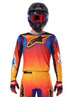 Alpinestars 2025 Fluid Wurx Jersey, wielokolorowa koszulka motocrossowa z pomarańczowo-czerwonym gradientem i fioletowymi akcentami, noszona z pasującym kompletem, czarny kask z niebiesko-czerwonymi goglami i czerwone rękawice, z dużym czarnym logo gwiazdy na piersi.