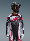 Koszulka Alpinestars 2026 Techstar Nomur, koszulka motocrossowa, czarno-biało-czerwona, noszona przez motocyklistę z dopasowanymi spodniami, czarnym kaskiem i białymi rękawicami, z dużym czerwonym logo Astars i nakrapianym wzorem graficznym, sportowy krój wyścigowy do rywalizacji w terenie