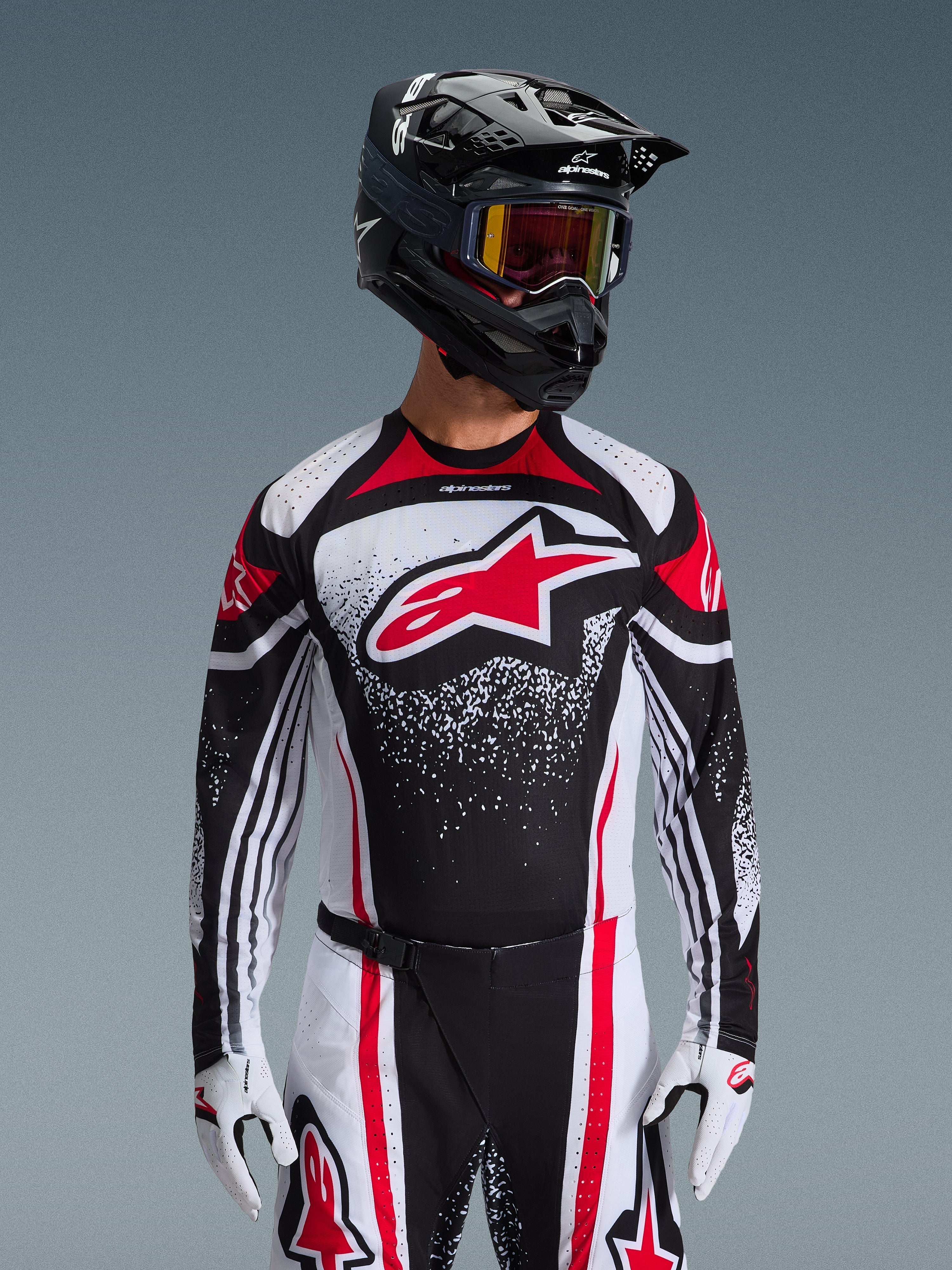 Koszulka Alpinestars 2026 Techstar Nomur, koszulka motocrossowa, czarno-biało-czerwona, noszona przez motocyklistę z dopasowanymi spodniami, czarnym kaskiem i białymi rękawicami, z dużym czerwonym logo Astars i nakrapianym wzorem graficznym, sportowy krój wyścigowy do rywalizacji w terenie