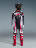 Kierowca motocrossowy ubrany w zestaw Alpinestars Techstar Nomur 2026, czarno-biało-czerwony, koszulka z dużym czerwonym logo gwiazdy i nakrapianym wzorem, pasujące spodnie z czerwonymi panelami bocznymi, czarny kask z przyciemnianymi goglami i czerwone buty motocrossowe.