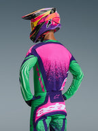 Motocyklista motocrossowy widziany od tyłu, ubrany w zestaw Alpinestars 2026 Techstar Nomur, koszulkę i pasujące spodnie w kolorach Deep Purple Green i Pink Fluo, z wyrazistymi grafikami i sportowym krojem wyścigowym, w zestawie z wielokolorowym kaskiem i żółtymi goglami.