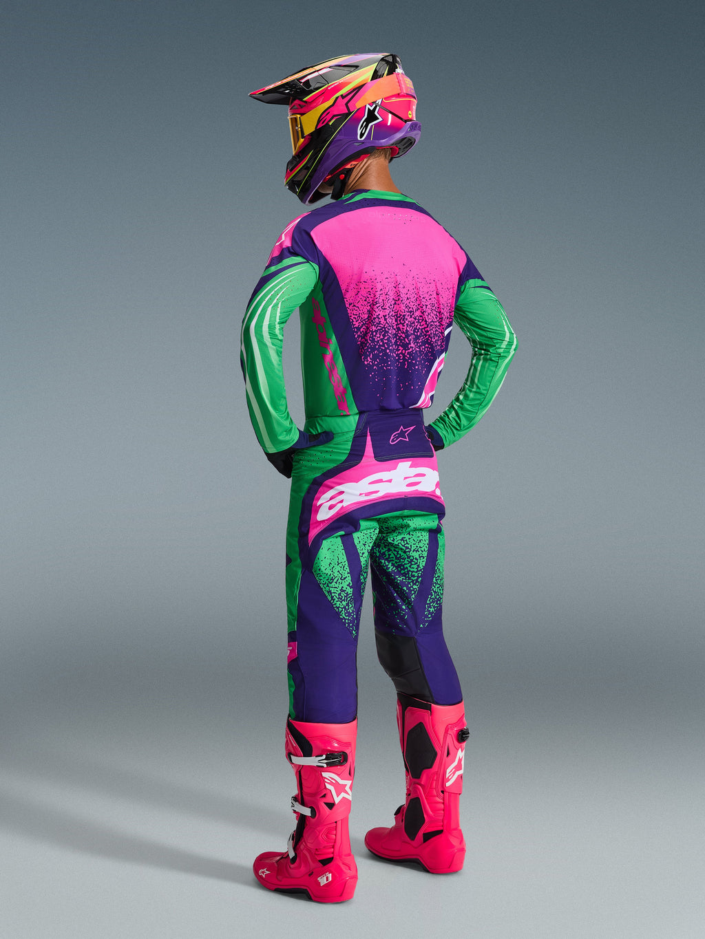 Motocyklista motocrossowy w zestawie Techstar Nomur 2026, w kolorach Deep Purple, Green i Pink Fluo, koszulka z różowym tyłem i nakrapianą teksturą, pasujące spodnie z białym logo, jaskrawe neonowe różowe buty i kolorowy kask, widok od tyłu całej sylwetki do wyścigów terenowych