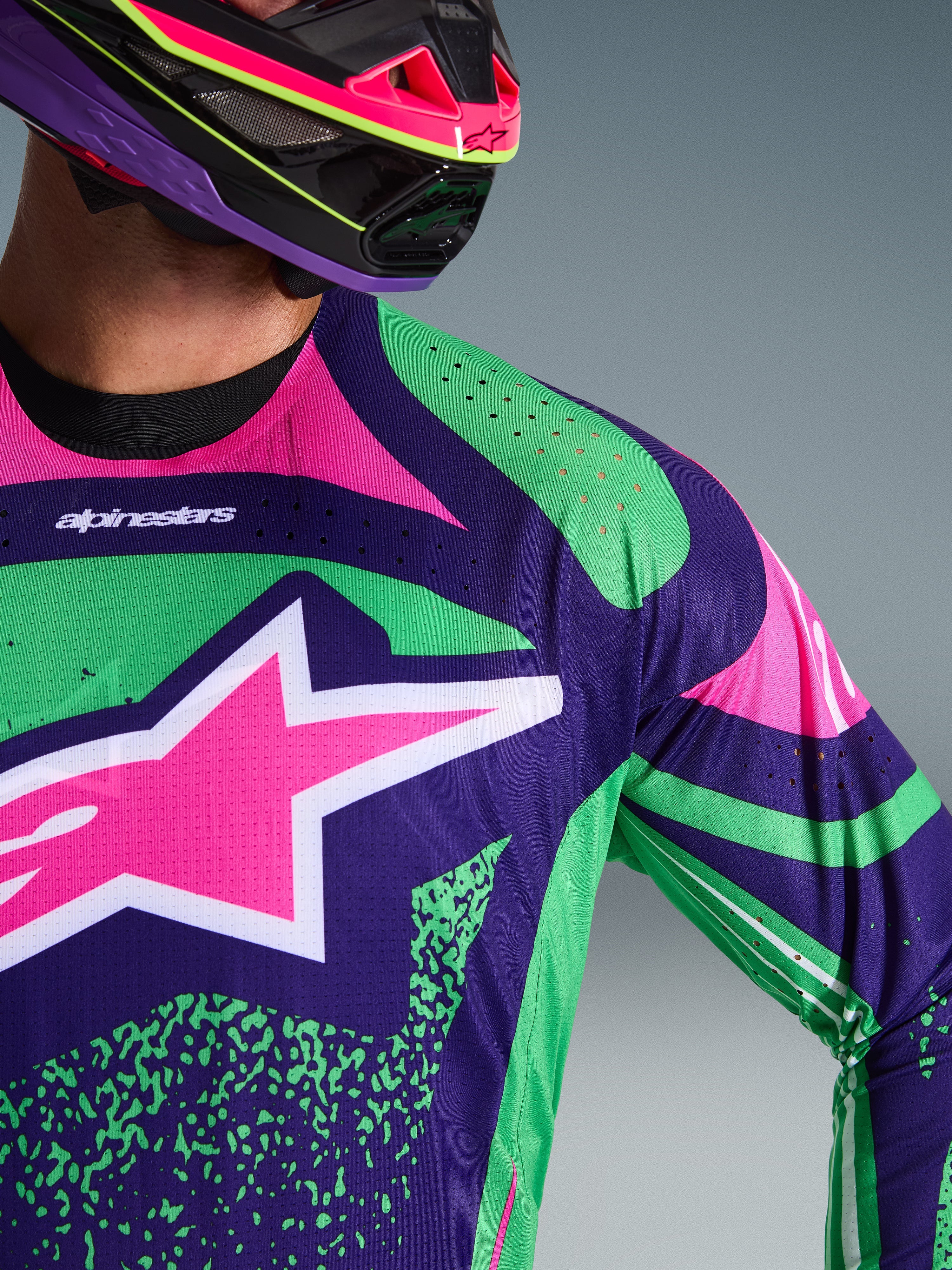 Koszulka Alpinestars Techstar Nomur 2026, zbliżenie na model w kolorze ciemnofioletowym z zielonymi i różowymi akcentami fluo, duże logo różowej gwiazdy na klatce piersiowej, laserowo wycinane perforacje na ramieniu, zawodnik w pasującym fioletowo-różowym kasku motocrossowym.