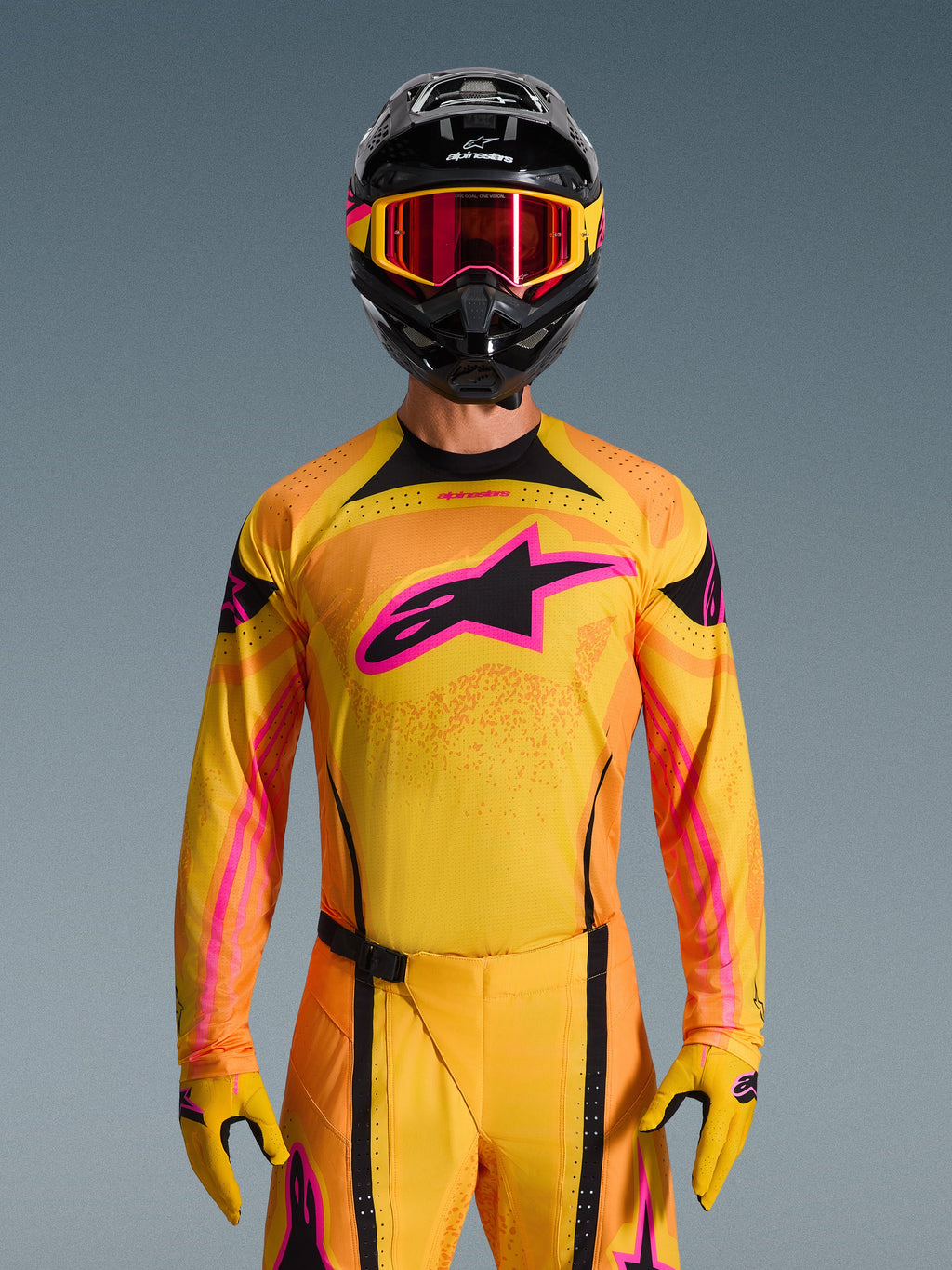 Koszulka Alpinestars Techstar Nomur 2026 w kolorze pomarańczowo-żółto-różowym, noszona przez motocyklistę motocrossowego w czarnym kasku i różowych goglach, z dużym różowym logo Alpinestars na piersi, sportowy krój wyścigowy zapewniający wydajność w terenie