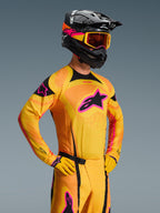 Alpinestars 2026 Techstar Nomur Jersey, koszulka motocrossowa, pomarańczowa, żółta i różowa, noszona przez motocyklistę w czarnym kasku i goglach, z dużym różowym logo gwiazdy na piersi, czarnymi akcentami i laserowo wycinanymi detalami wentylacyjnymi.