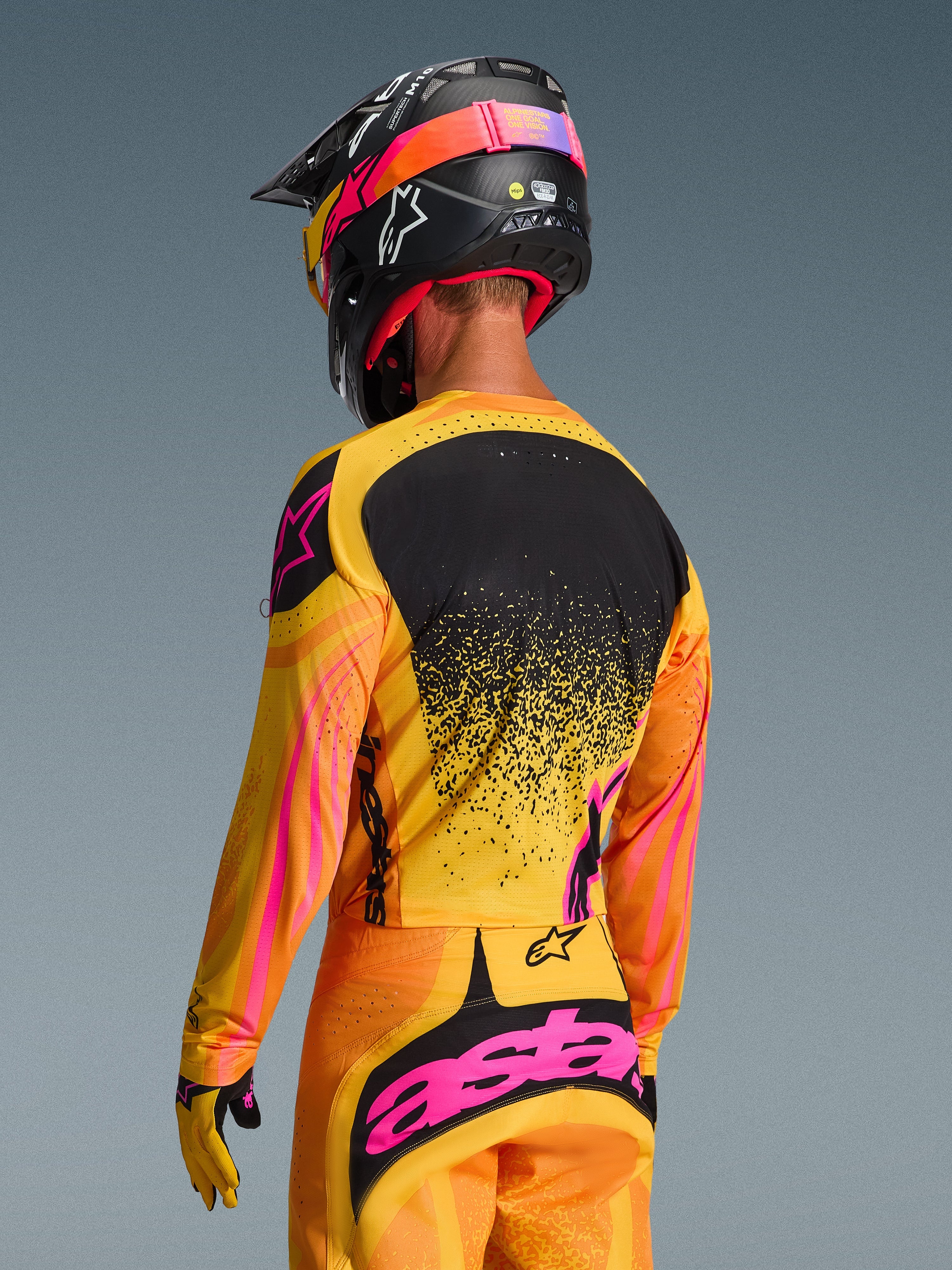 Alpinestars 2026 Techstar Nomur Jersey w kolorze pomarańczowo-żółto-różowym, widok z tyłu zawodnika motocrossowego w pełnym stroju z pasującymi spodniami i czarnym kaskiem, jaskrawy design z żółtymi i różowymi akcentami oraz czarnym panelem na plecach, sportowy krój wyścigowy.