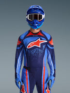 Alpinestars 2026 Techstar Nomur Jersey, koszulka motocrossowa, ciemnogranatowa, pomarańczowa i niebieska UCLA, noszona z pasującymi spodniami, niebieskim kaskiem i rękawicami, z białym logo gwiazdy na piersi, dynamicznymi liniami i kropkowanymi wzorami