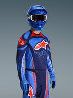 Zawodnik motocrossowy w koszulce Alpinestars 2026 Techstar Nomur, strój wyścigowy w barwach granatowo-pomarańczowych i błękitnych (UCLA Blue), z dużym pomarańczowym logo Astars, pasujący niebieski kask i rękawice, sportowy ubiór do zawodów terenowych