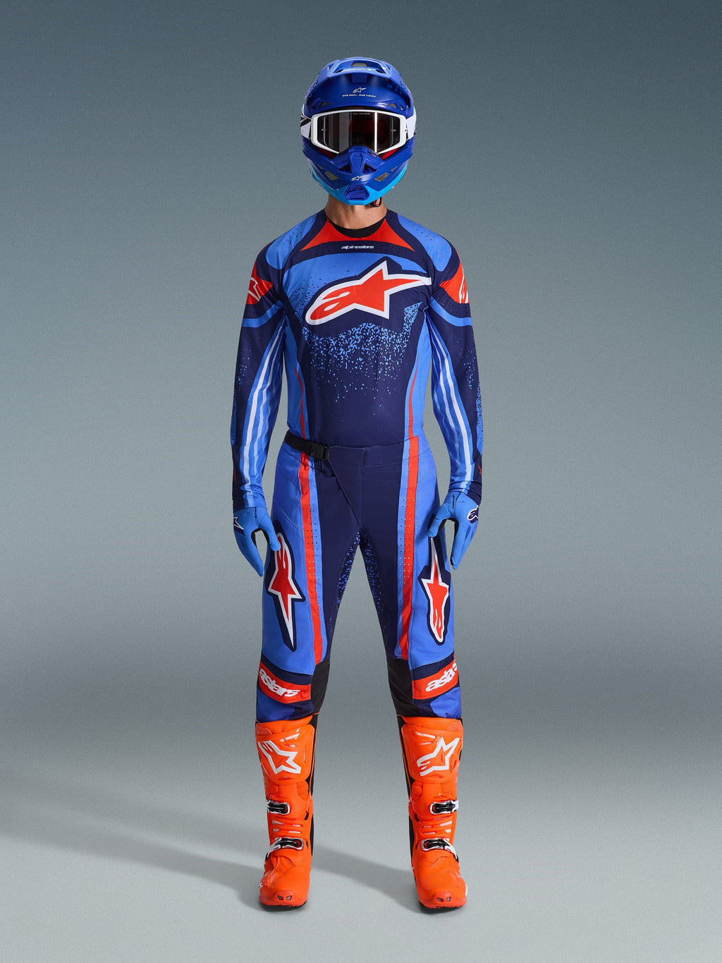 Zawodnik motocrossowy ubrany w koszulkę Alpinestars 2026 Techstar Nomur w kolorach granatowym, pomarańczowym i błękitnym, w zestawie z dopasowanymi spodniami, niebieskim kaskiem z goglami oraz jaskrawopomarańczowymi butami wyścigowymi, kompletny profesjonalny strój off-road