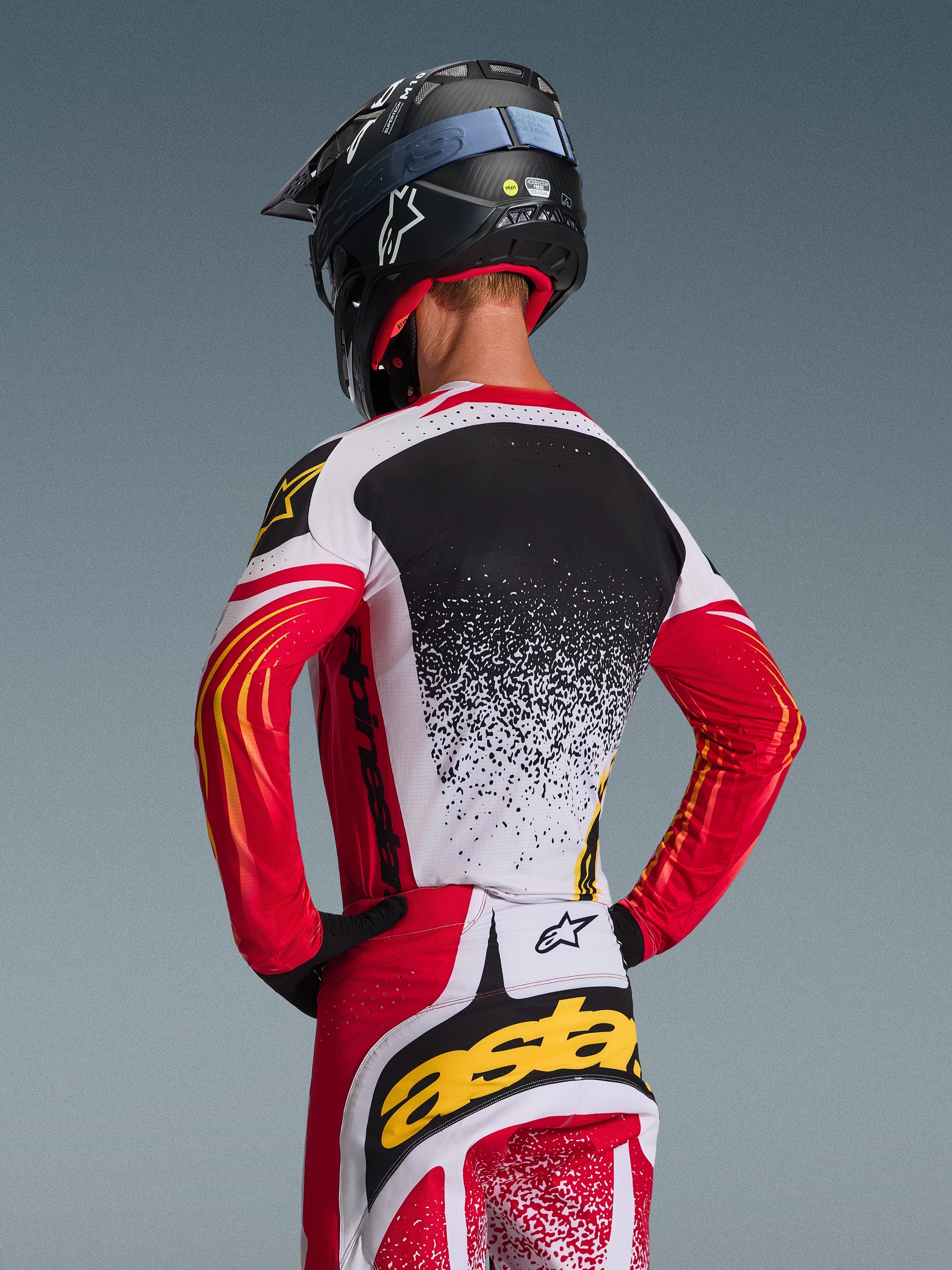 Motocyklista motocrossowy widziany od tyłu w koszulce Alpinestars 2026 Techstar Nomur i dopasowanych spodniach, czerwono-biało-czarny wzór z żółtymi akcentami i nakrapianym gradientem na plecach, czarny kask Alpinestars i czarne rękawice, profesjonalny zestaw odzieży do wyścigów off-road