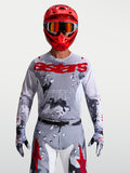 Alpinestars Koszulka Racer Air Lone, męska koszulka motocrossowa z oddychającej siatki, wzór szary kamuflaż i czerwony, ergonomiczny krój wyścigowy, na modelu w kasku i rękawicach.