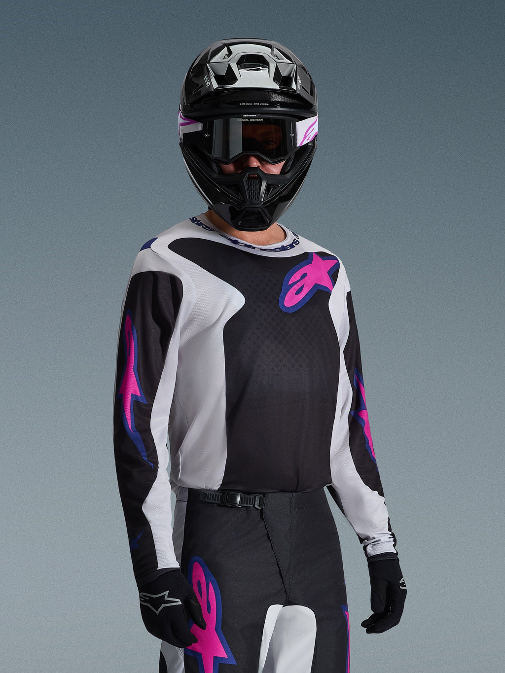 Motocyklista motocrossowy w koszulce Alpinestars 2026 Fluid Grid, czarna, jasnoszara i fioletowa, duże różowe i fioletowe logo gwiazdy, dopasowane spodnie, czarny kask i gogle, kompletny zestaw wyścigowy off-road