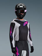 Motocyklista motocrossowy w koszulce Alpinestars 2026 Fluid Grid, czarna, jasnoszara i fioletowa, duże różowe i fioletowe logo gwiazdy, dopasowane spodnie, czarny kask i gogle, kompletny zestaw wyścigowy off-road