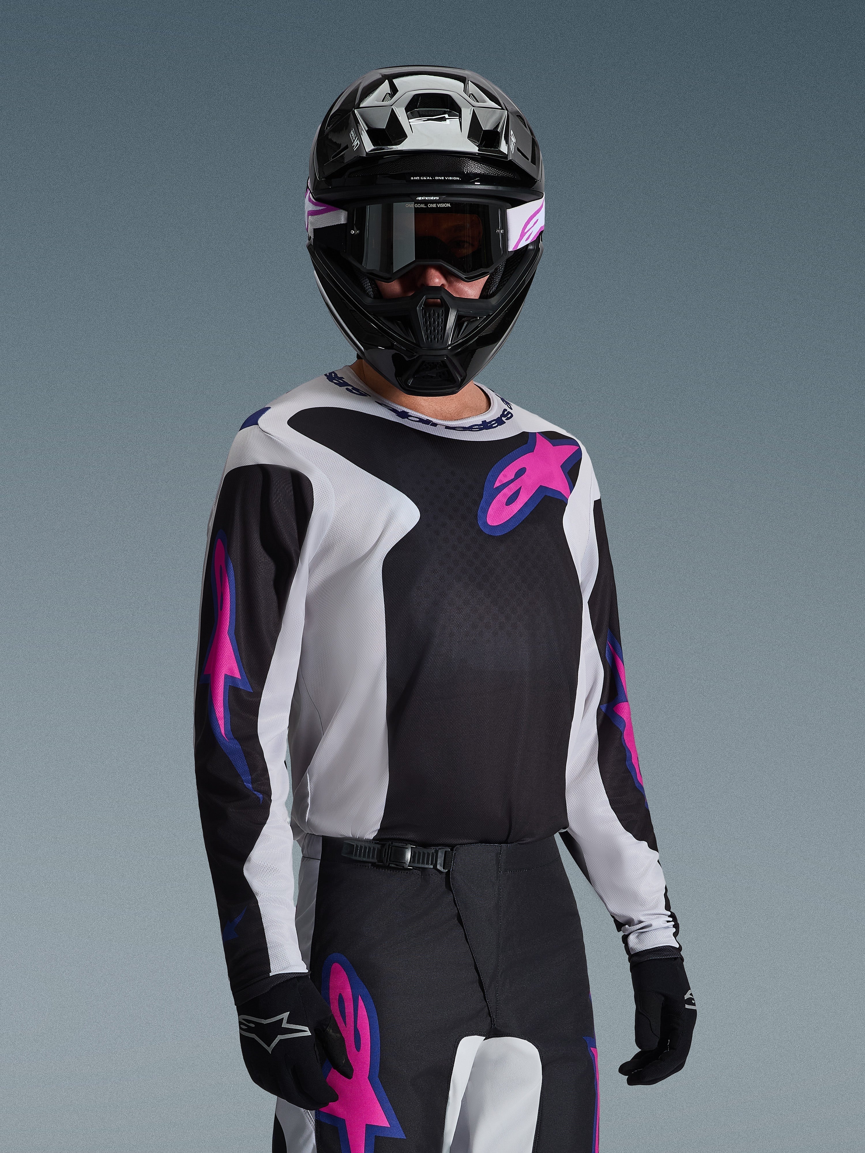 Motocyklista motocrossowy w koszulce Alpinestars 2026 Fluid Grid, czarna, jasnoszara i fioletowa, duże różowe i fioletowe logo gwiazdy, dopasowane spodnie, czarny kask i gogle, kompletny zestaw wyścigowy off-road