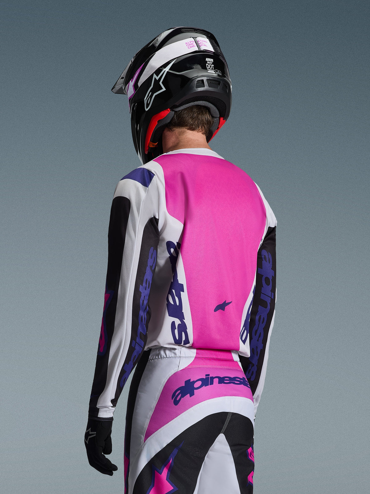 Motocyklista motocrossowy widziany od tyłu w koszulce Alpinestars 2026 Fluid Grid w kolorze czarnym, białym i fioletowym, pasujących spodniach z wyraźnym brandingiem, czarnym kasku z różowymi akcentami i czarnych rękawicach; kompletny, spójny zestaw odzieży off-road.