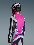 Motocyklista motocrossowy widziany od tyłu w koszulce Alpinestars 2026 Fluid Grid w kolorze czarnym, białym i fioletowym, pasujących spodniach z wyraźnym brandingiem, czarnym kasku z różowymi akcentami i czarnych rękawicach; kompletny, spójny zestaw odzieży off-road.