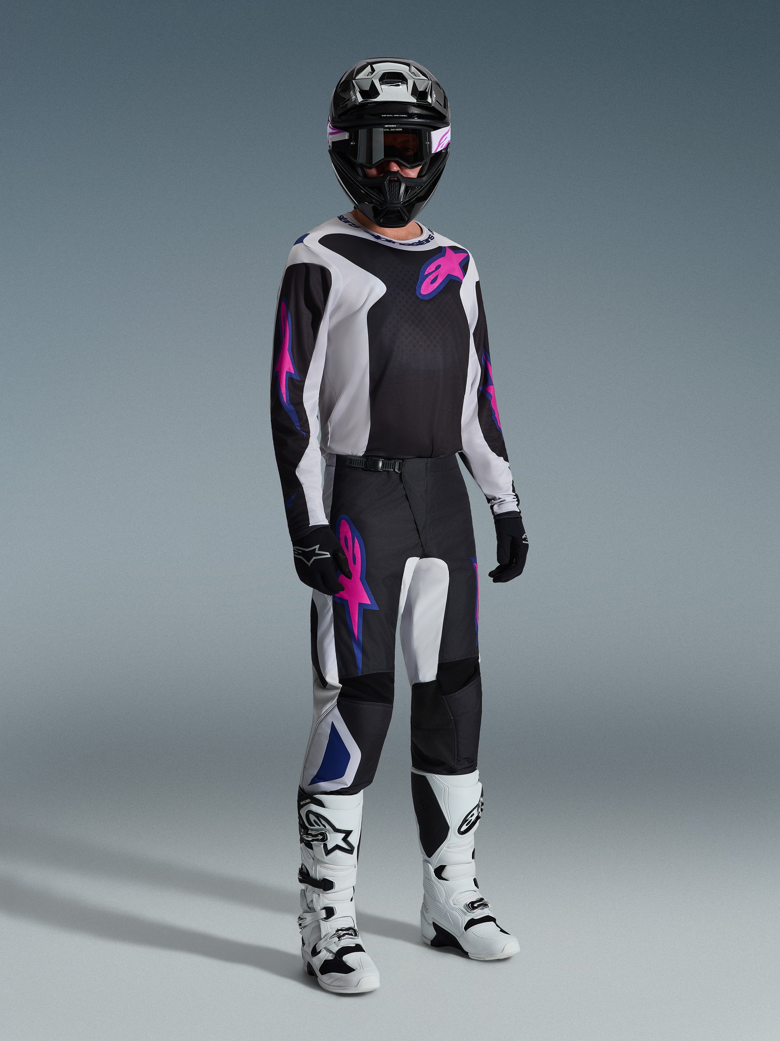 Alpinestars 2026 Fluid Grid Gear Set, motocyklista motocrossowy w czarno-białej bluzie i spodniach z różowymi i niebieskimi akcentami, czarny kask, czarne rękawice i białe buty, kompletny zestaw wyścigowy na szarym tle