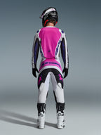 Koszulka i spodnie Alpinestars Fluid Grid 2026, zestaw odzieży motocrossowej, różowy, biały i fioletowy, widok z tyłu prezentujący ergonomiczny sportowy krój, zestawiony z czarnym kaskiem i białymi butami, zaprojektowany pod kątem wydajności terenowej.