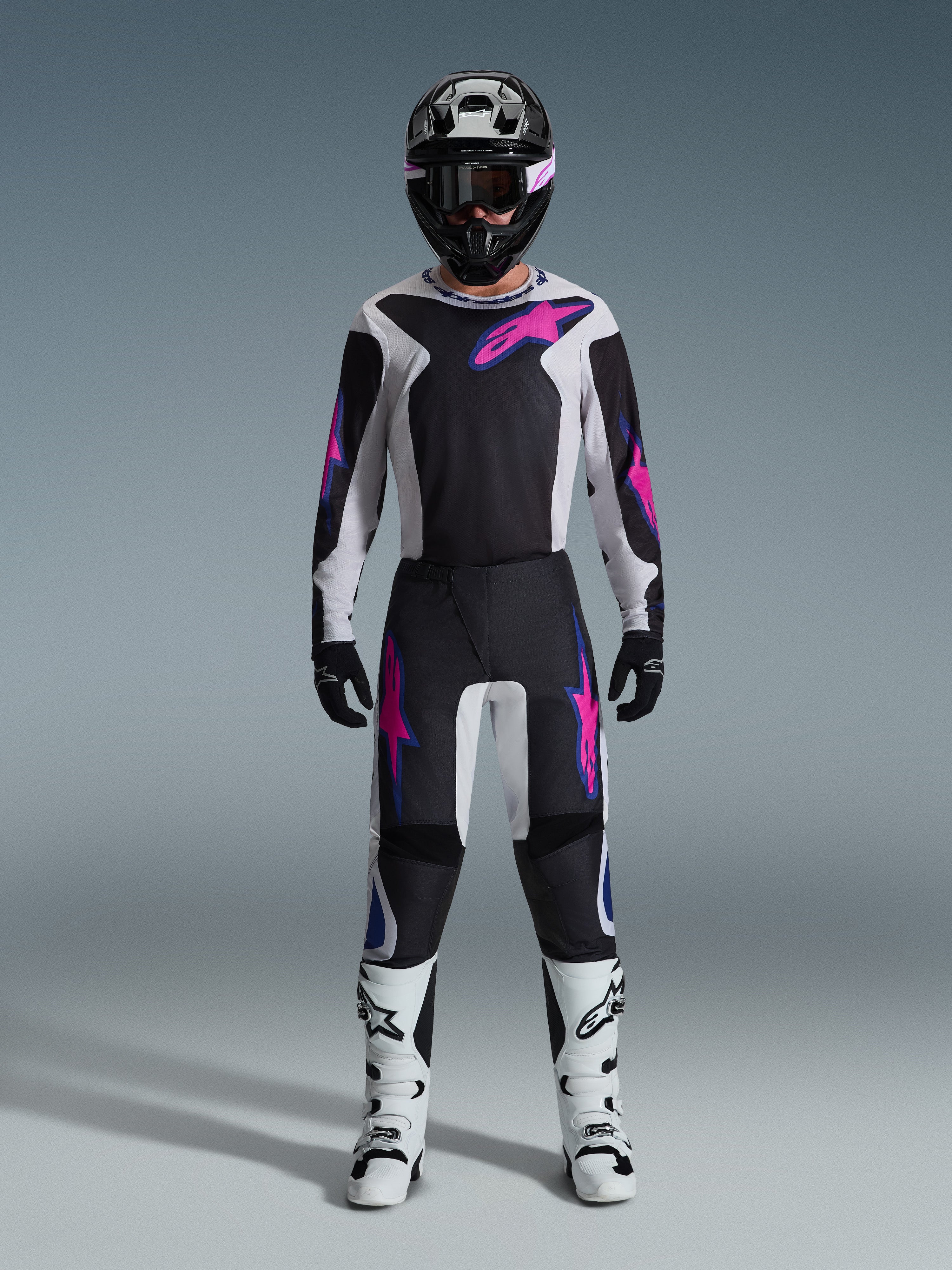 Motocyklista motocrossowy w pełnym stroju wyścigowym, koszulka Alpinestars 2026 Fluid Grid i pasujące spodnie w kolorze czarnym i jasnoszarym z wyrazistymi fioletowymi akcentami, czarny kask z fioletowymi goglami, białe buty motocrossowe i czarne rękawice, skoordynowany zestaw do jazdy off-road