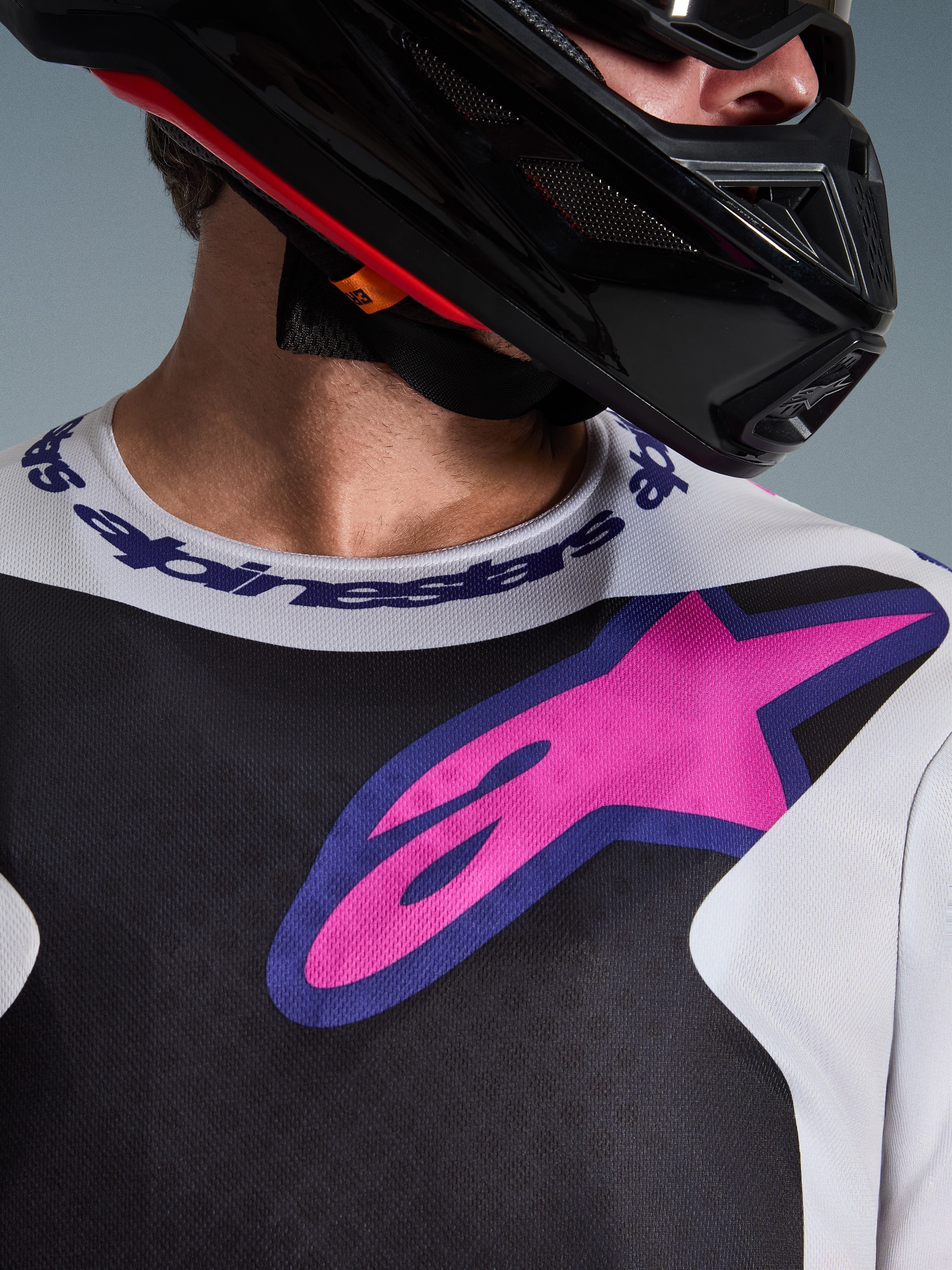 Zbliżenie na koszulkę Alpinestars 2026 Fluid Grid, koszulka motocrossowa, czarna, jasnoszara i fioletowa, z wyrazistym różowo-fioletowym logo gwiazdy na piersi i markowym sportowym kołnierzykiem, noszona z czarnym kaskiem motocrossowym.