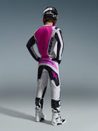 Alpinestars 2026 Fluid Grid Gear Set, zawodnik motocrossowy widziany od tyłu, biała bluza z różowym panelem na plecach i fioletowym logo, dopasowane biało-czarne spodnie z różowymi akcentami, biało-czarne buty i czarny kask, skoordynowany strój do wyścigów terenowych