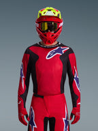 Zawodnik motocrossowy w zestawie Alpinestars 2026 Fluid Grid, czerwona koszulka z czarnymi panelami bocznymi i białym logo gwiazdy, pasujące czerwone spodnie i rękawice, czerwono-żółty kask ze złotymi goglami, wysokowydajny sprzęt wyścigowy do jazdy off-road