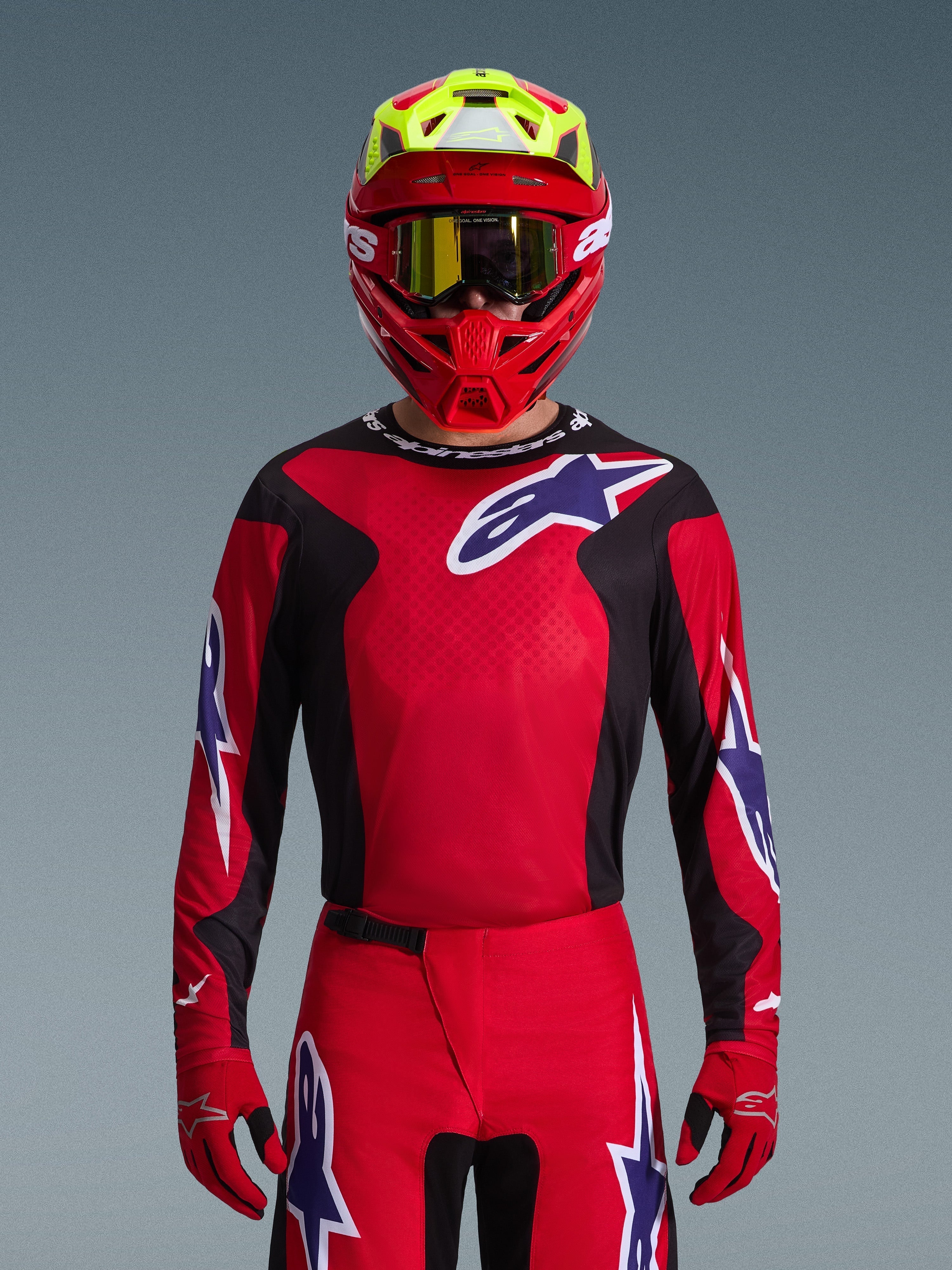 Koszulka Alpinestars 2026 Fluid Grid, koszulka motocrossowa, czerwono-czarno-fioletowa, noszona przez motocyklistę w czerwonym i neonowożółtym kasku oraz złotych goglach, z dużym logo gwiazdy na klatce piersiowej i rękawach, dopasowany zestaw do wyścigów off-road