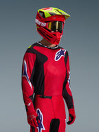 Zawodnik motocrossowy w zestawie Alpinestars 2026 Fluid Grid, czerwono-czarna koszulka z białymi i niebieskimi akcentami logo, pasujące czerwono-czarne spodnie, czerwony kask z żółtymi detalami i złotymi goglami, dopasowany strój do wyścigów terenowych