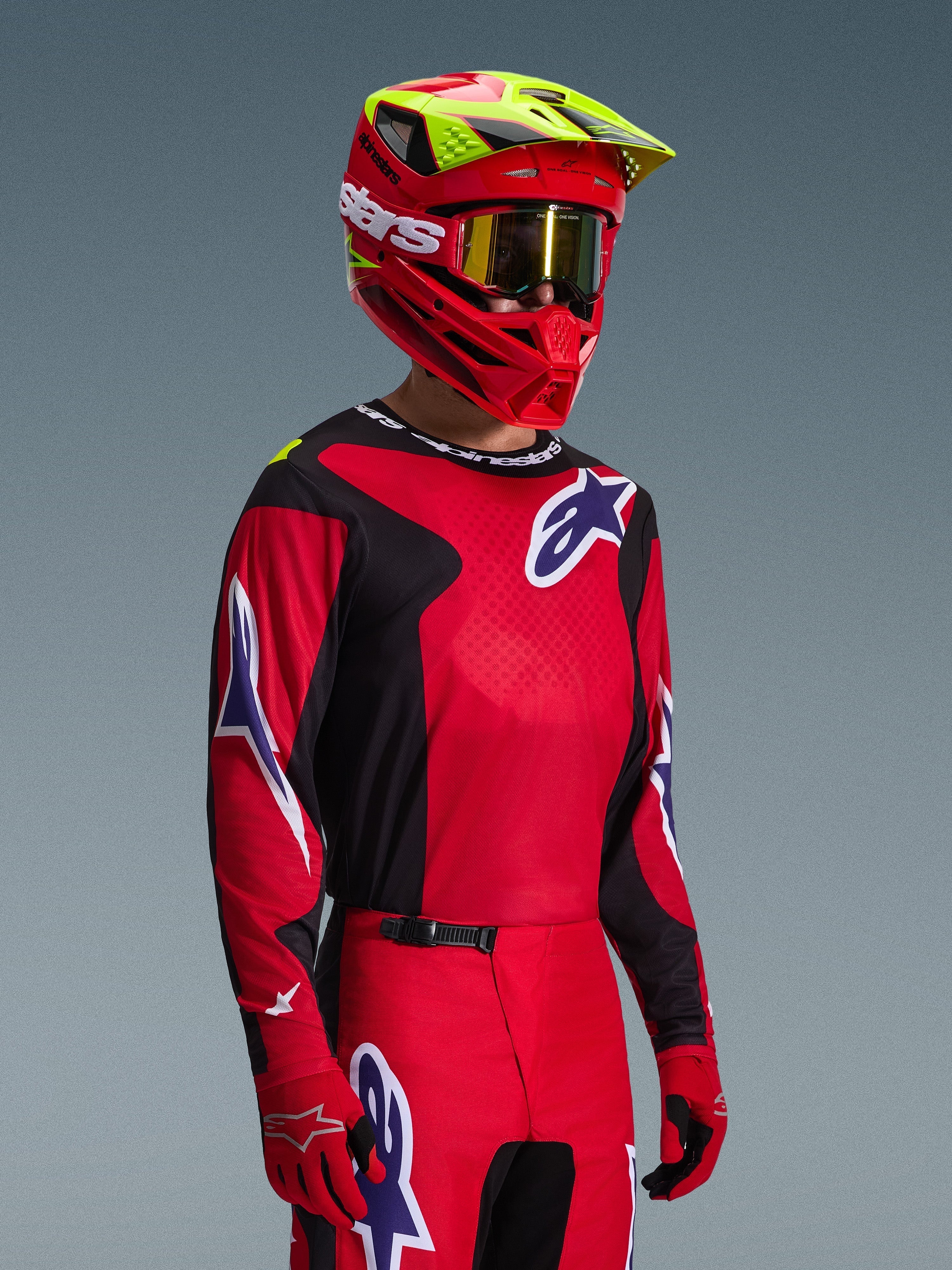 Zawodnik motocrossowy w zestawie Alpinestars 2026 Fluid Grid, czerwono-czarna koszulka z białymi i niebieskimi akcentami logo, pasujące czerwono-czarne spodnie, czerwony kask z żółtymi detalami i złotymi goglami, dopasowany strój do wyścigów terenowych