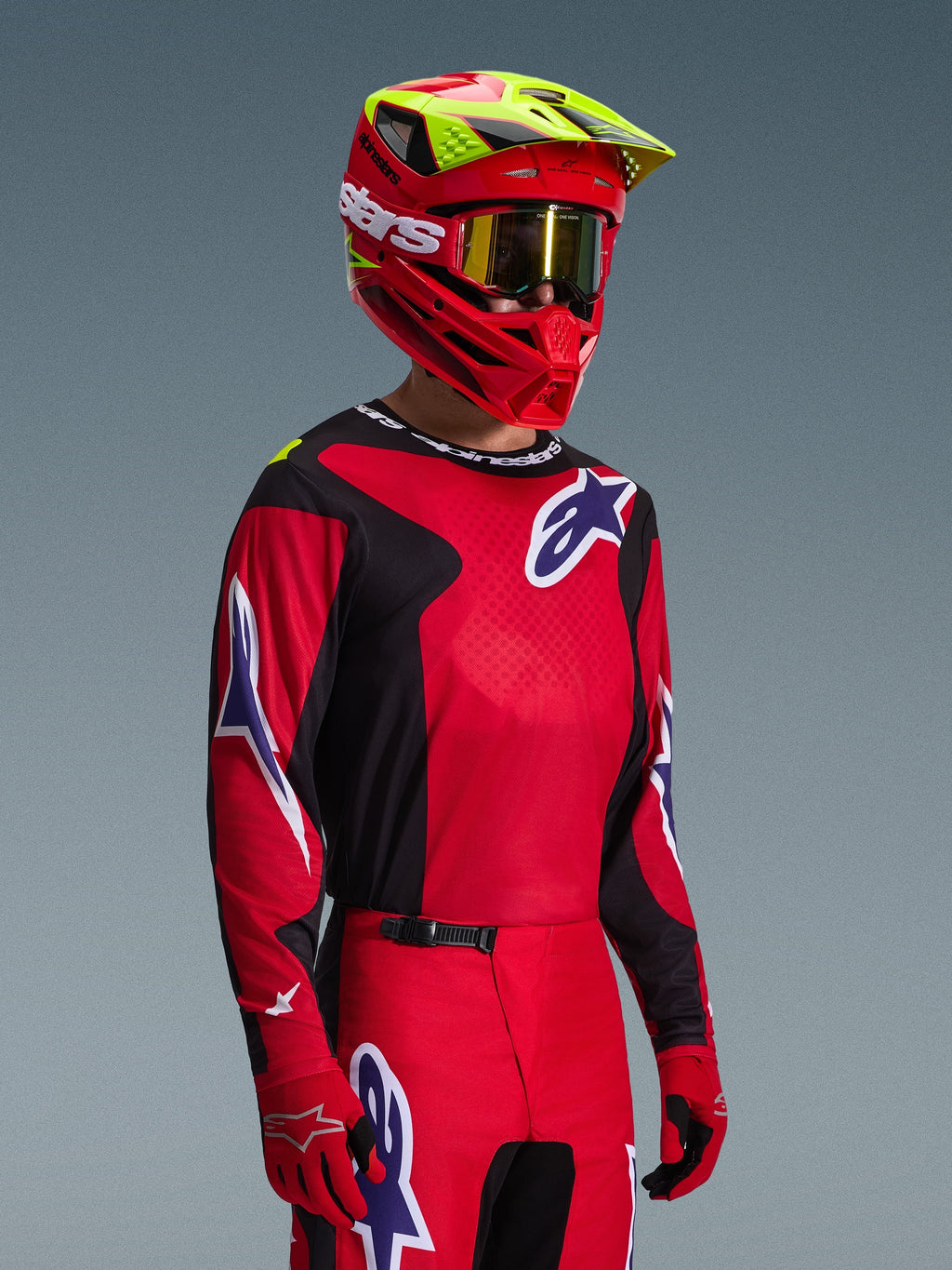 Motocyklista motocrossowy ubrany w koszulkę Alpinestars 2026 Fluid Grid w kolorze czerwonym, czarnym i fioletowym, zestawioną z dopasowanymi spodniami motocrossowymi, czerwonym kaskiem z neonowożółtymi akcentami, złotymi goglami i czerwonymi rękawicami, kompletny strój wyścigowy off-road.