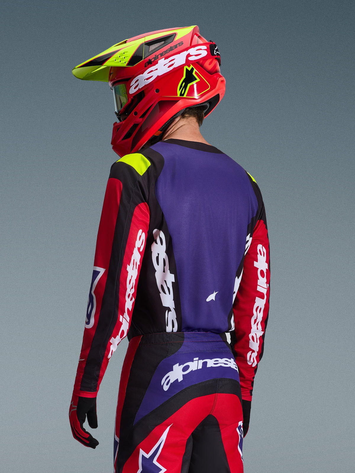 Motocyklista motocrossowy ubrany w koszulkę Alpinestars 2026 Fluid Grid, czerwono-czarno-fioletową, widok z tyłu pokazujący fioletowy panel na plecach i czerwone rękawy z białym logo, dopasowane spodnie oraz czerwony i neonowo-żółty kask, spójny zestaw do wyścigów off-road