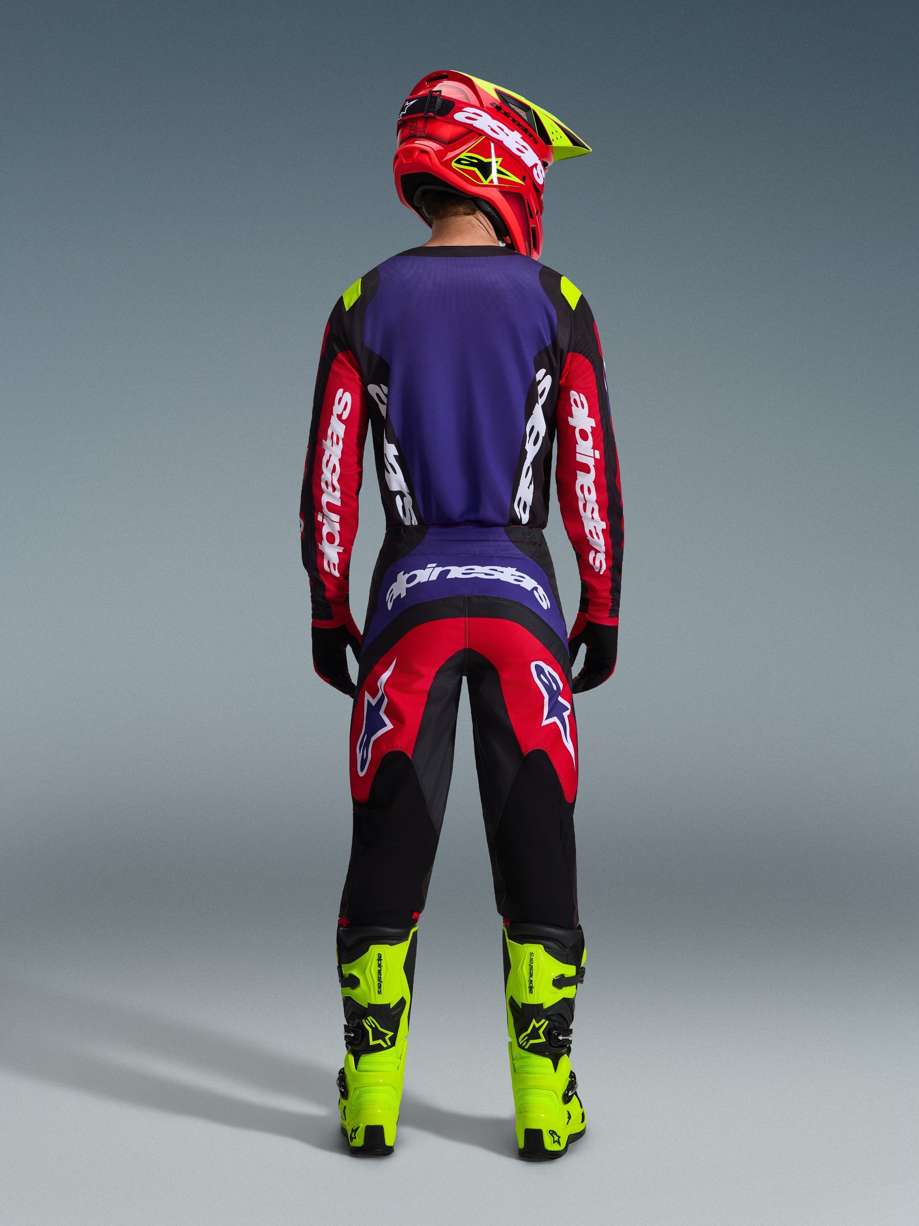 Zawodnik motocrossowy widoczny od tyłu w koszulce 2026 Fluid Grid w kolorze Red Black Purple, białe logo Alpinestars na długich rękawach, pasujące spodnie motocrossowe, neonowożółte buty i czerwono-żółty kask, kompletny zestaw do wyścigów off-road