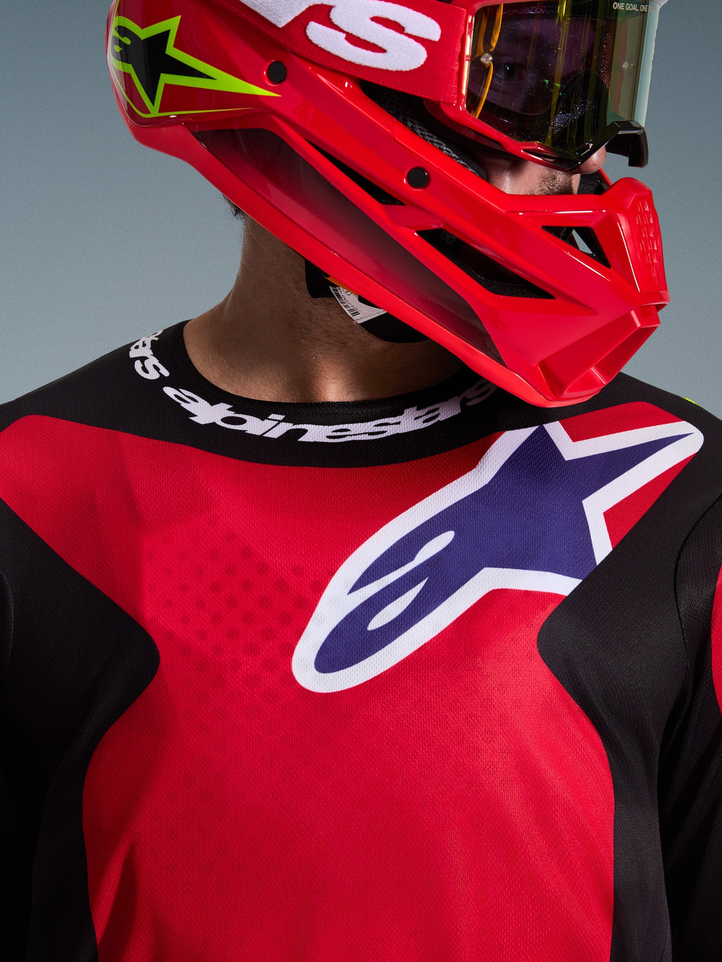 Alpinestars 2026 Fluid Grid Jersey, koszulka motocrossowa w kolorach czerwonym, czarnym i fioletowym, zbliżenie na zawodnika, duże logo na piersi, logowany sportowy kołnierzyk, czerwony kask i gogle, techniczna tkanina poliestrowa i sportowy krój.