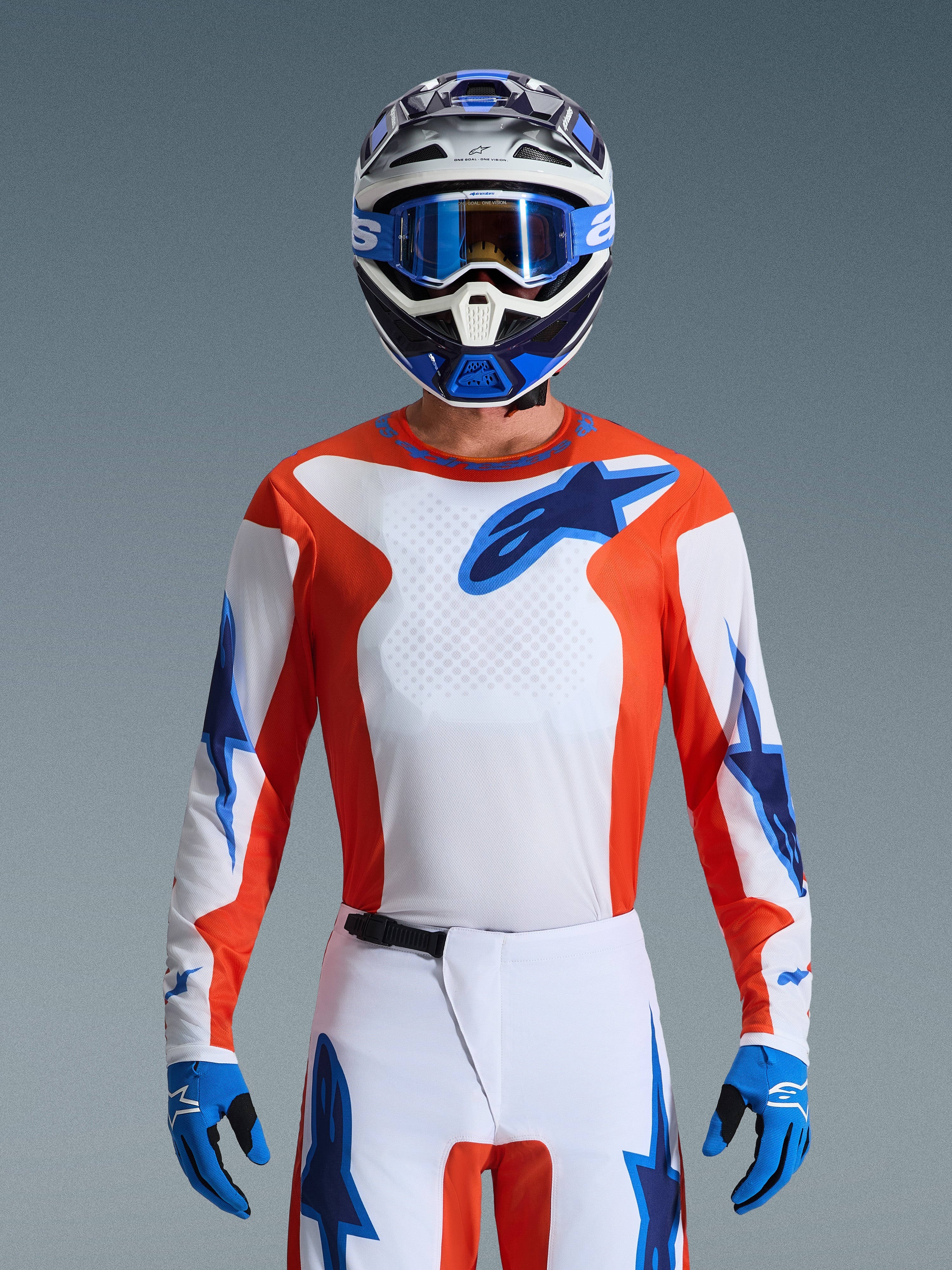 Motocyklista motocrossowy w koszulce Fluid Grid Jersey 2026 w kolorze Orange Ucla Blue, biały panel piersiowy z pomarańczowymi panelami bocznymi i niebieskim logo Alpinestars, biało-niebieskie spodnie, niebieskie rękawice i biało-niebieski kask, profesjonalny wygląd off-road